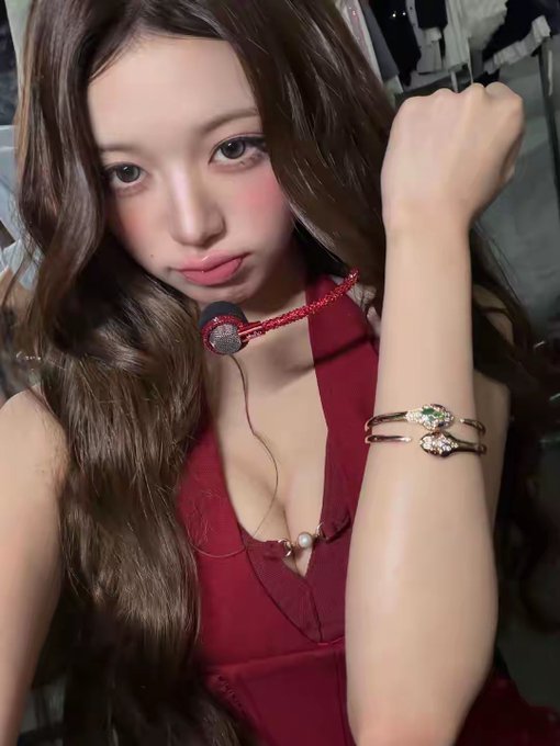 A young woman with long brown hair poses in a selfie, wearing a deep red halter dress, a snake-shaped brooch, and a gold snake bracelet on her wrist.