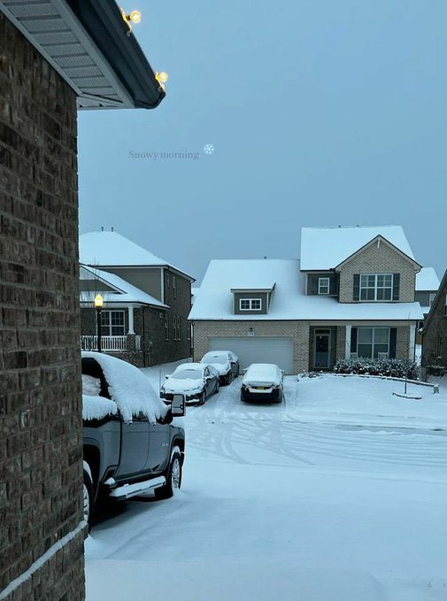 The image shows a snowy street with several cars parked along the side of the road. The snow-covered houses are visible behind the parked cars, creating an overall winter scene. A car covered in snow can be seen near one of the houses, adding to the wintry atmosphere.