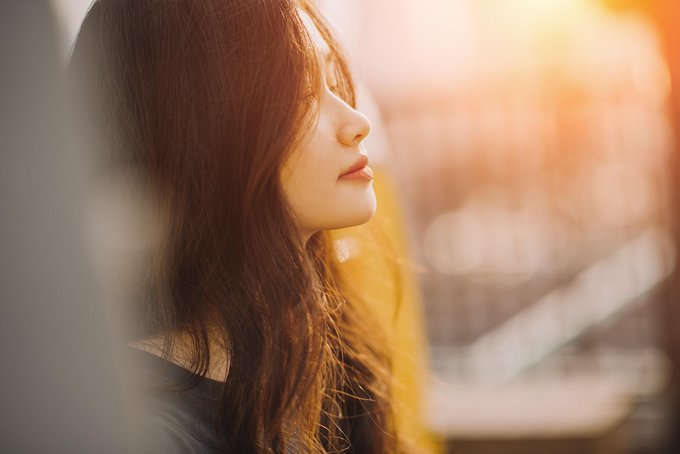 A side profile portrait of a young woman with long brown hair, her eyes closed as warm, golden sunlight streams in from the right, creating a soft bokeh effect in the background.