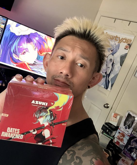 A man with blonde and black hair holds up an AZUKI trading card game box labeled 'GATES AWAKENED' in front of a monitor displaying anime artwork.