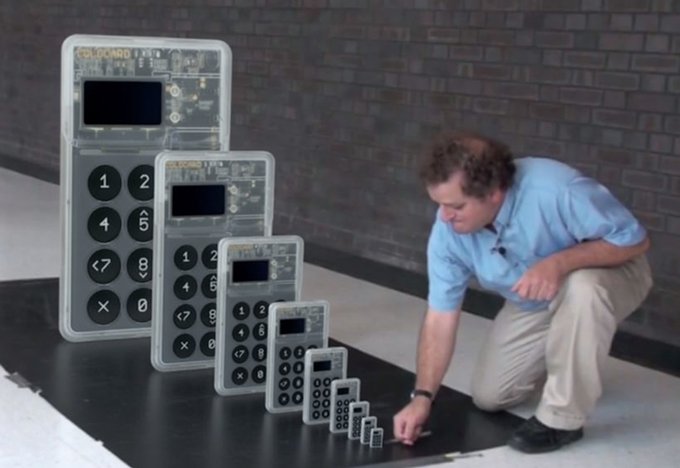A man kneels beside a line of calculators arranged in descending size, showcasing the dramatic scale difference between the largest and smallest models. The caption playfully suggests these nested devices, like Russian dolls, reveal "the evolution of calculators"—from the massive ColdCard-sized unit down to a tiny button-sized version—hinting at the novelty-sized ColdCard hardware wallet prize.