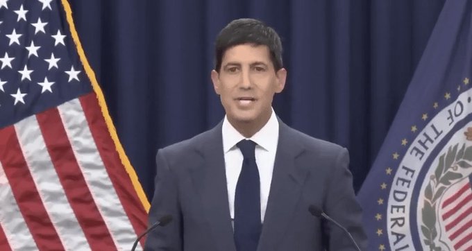 A man in a dark suit and blue tie speaking at a podium, with an American flag on the left and a Federal Reserve seal on the right in the background.