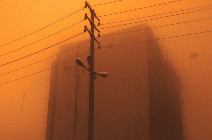 Silhouettes of buildings and a street lamp against a hazy, orange-tinted sky, with power lines stretching across the frame.