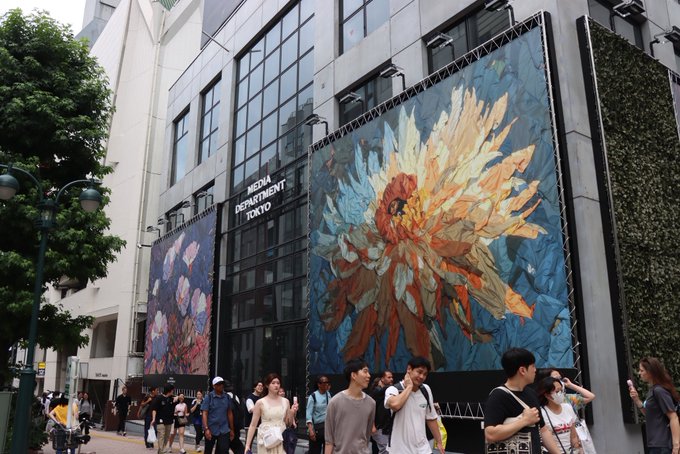 Crowds of pedestrians walk past a building labeled "MEDIA DEPARTMENT TOKYO," which features large, vibrant floral murals on its exterior walls, including a prominent sun-like orange and yellow bloom on a blue background. A cyclist rides along the street as people go about their day in this busy urban scene.