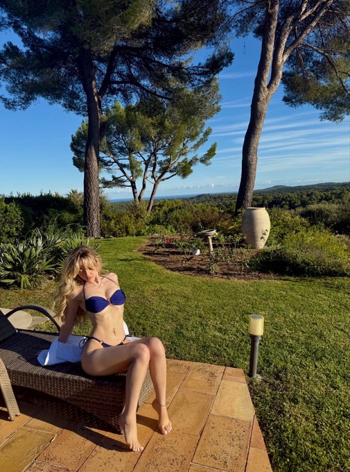 A blonde woman in a blue bikini sitting on a wicker lounge chair on a stone patio, with a lush green lawn, large trees, and an ocean view in the background.