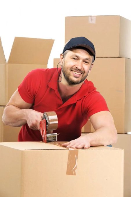 A smiling man in a red polo shirt and dark cap is taping a cardboard box with a tape dispenser, surrounded by stacked cardboard boxes.
