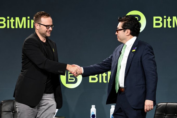 Two men in suits shake hands on stage at what appears to be a BitMine event, with one man wearing a green tie and glasses and the other in a black blazer.