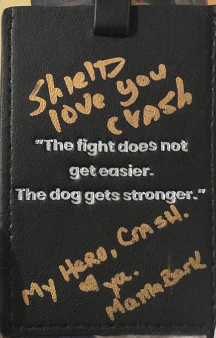 A black leather item with gold handwritten text 'Sheild love you Crash' and white printed text reading, 'The fight does not get easier. The dog gets stronger,' followed by more handwritten text 'My Hero, Crash. Ya, Mama Bear.'