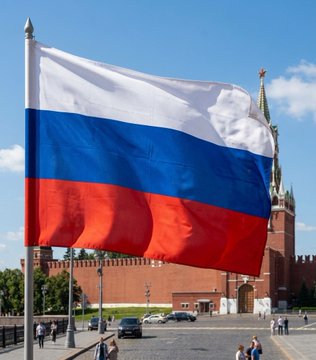 The image features a large Russian flag flying high above a city street with people walking around. The flag has red, white, and blue colors, which are traditional for Russia. In the background, there's a tall building that appears to be a castle-like structure, possibly representing the Kremlin in Moscow. A post on social media is visible, discussing Bitcoin and Ethereum as investment options. This post seems to be promoting Bitcoin or Ethereum as an alternative to traditional Russian currency, which could potentially impact the local economy and financial landscape of Russia.

There are several people walking around the street, some carrying backpacks and handbags. A few cars can also be seen parked along the street. The overall atmosphere suggests a bustling city with diverse interests in technology and finance.