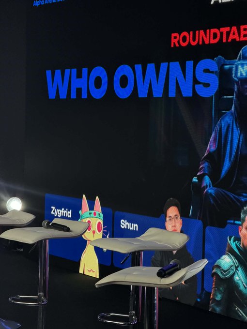 The image shows a Coinfest Asia roundtable stage with the banner text "WHO OWNS" prominently displayed in blue, alongside stool name placards featuring a yellow cat mascot ("Zygfrid") and a profile labeled "Shun." The scene aligns with the post about attending the Coinfest Asia roundtable while showcasing the @cetsongold PFP, capturing the conference setting where the crypto discussion took place.