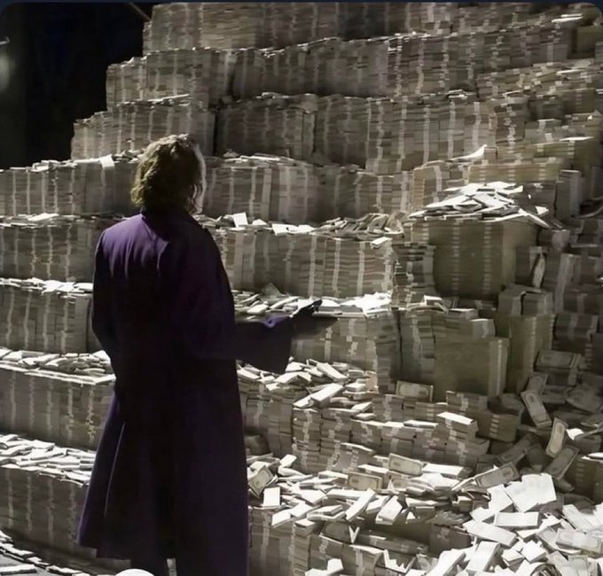 A person with shoulder-length hair, wearing a long purple coat, stands facing a massive pile of stacked banded currency.