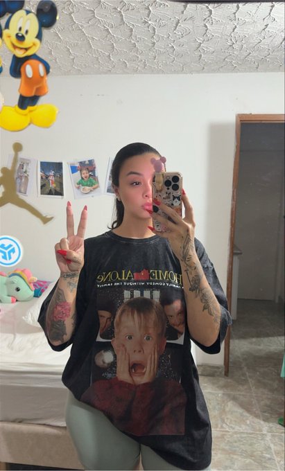 A mirror selfie of a woman with dark hair and tattoos making a peace sign, wearing an oversized black "HOME ALONE" t-shirt featuring the movie's iconic child character, with a Mickey Mouse balloon visible in the background.