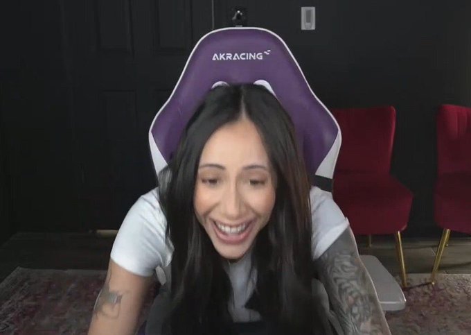 A woman with long dark hair and tattooed arms laughs while seated in front of a purple and white "AKRACING" gaming chair, with two red velvet chairs visible behind her. The caption and post suggest she's reacting humorously to another crypto hack.