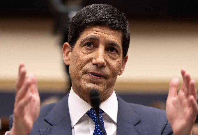 A man in a dark suit and blue patterned tie speaks while gesturing with both hands raised, appearing to testify before a microphone.
