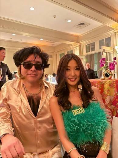 A man wearing sunglasses and a gold satin shirt poses alongside a woman in a green feathered dress at what appears to be a formal event.