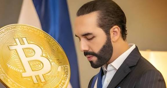 A man in a dark suit stands in front of a blue and white flag, with a large golden Bitcoin coin overlay displayed on the left side of the image.