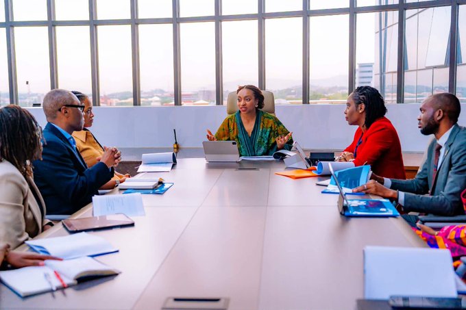 The image shows a group of people sitting around a conference table, engaged in discussion or meeting. There are at least six individuals visible in the scene, including a woman seated at the head of the table who appears to be leading the conversation. The table is covered with various items such as laptops and books, suggesting that they might be working on a project or discussing important matters related to their work.