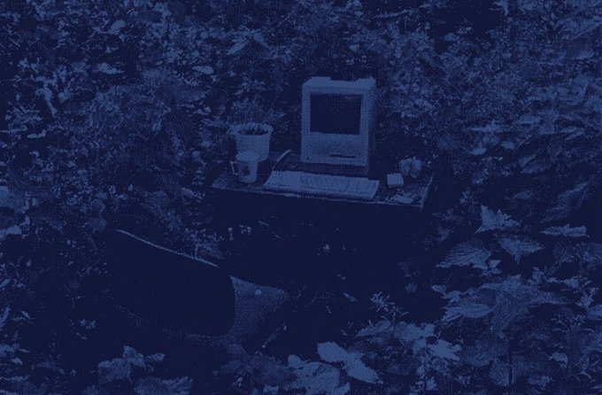 The image features a computer monitor sitting on top of a desk, surrounded by various objects such as a keyboard, mouse, cup, and chair. The scene appears to be set in an outdoor area with trees and bushes surrounding the workspace. A caption below the image reads "Someone aped $Surplus before I got home. Gg." This suggests that someone has been using their computer for an extended period of time or perhaps has not had the chance to clean up after themselves, resulting in a cluttered desk environment.