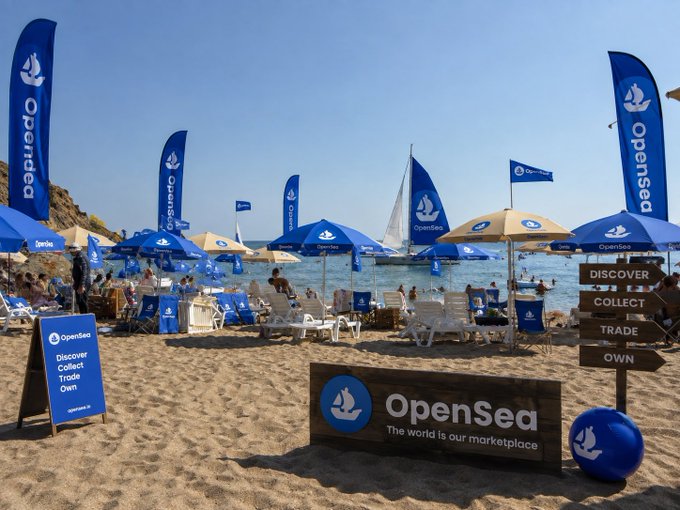 An OpenSea-branded beach setup with blue flags, umbrellas, and signs reading 'Discover Collect Trade Own', with the tagline 'The world is our marketplace'.