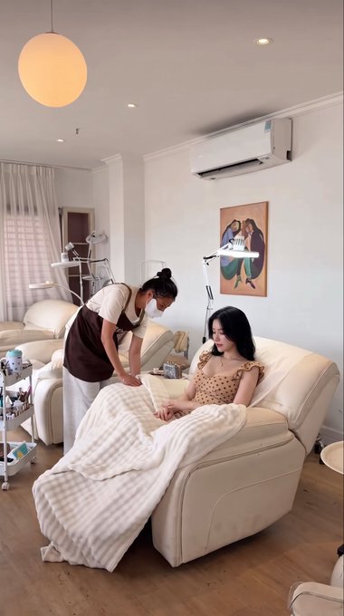 Inside a brightly lit treatment room, an esthetician in a brown apron attends to a client reclining on a cream-colored spa bed covered with a striped blanket.