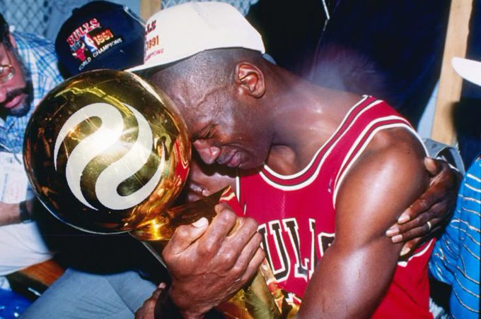 The image features a man wearing a red jersey, holding a trophy with both hands while hugging another person who is also wearing a red jersey. The two people are standing close together, and there's an additional person partially visible behind them. A sports ball can be seen in the background as well.