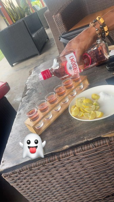 A hand pours Smirnoff vodka into a row of shot glasses on a wooden holder, with a plate of lime wedges nearby on a wicker table, accompanied by a cartoon ghost emoji.