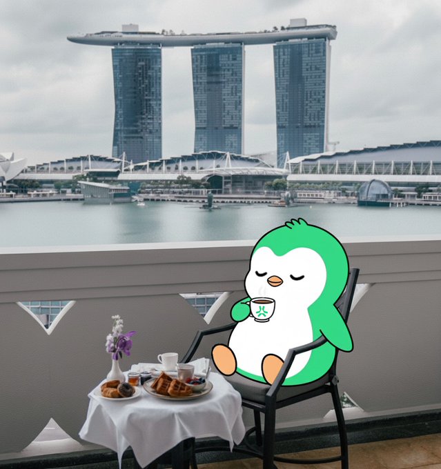 A green cartoon penguin holding a teacup sitting at a small table with breakfast food, with the Marina Bay Sands hotel and waterfront visible in the background of Singapore.