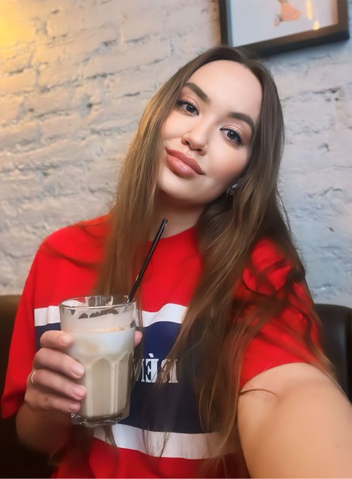 A young woman with long brown hair posing for a selfie while holding a glass of iced coffee with a black straw, wearing a red and white shirt.