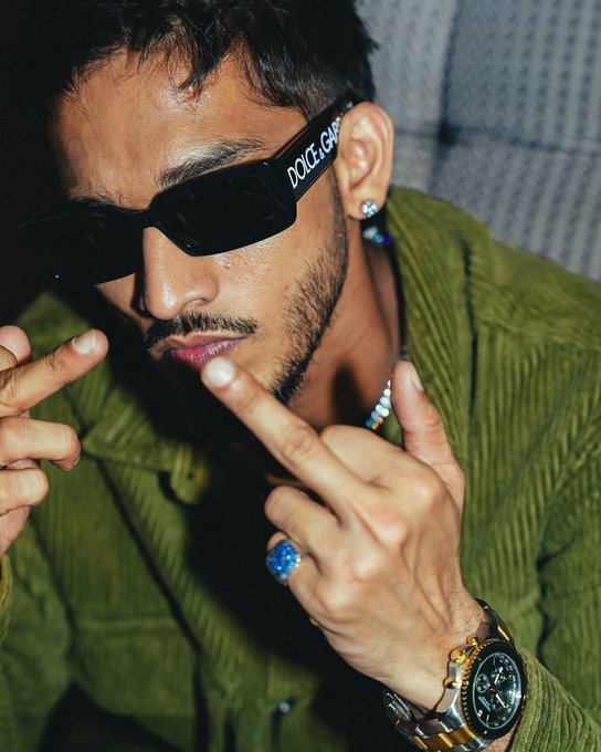 A close-up photo of a man wearing 'DOLCE & GABBANA' sunglasses, a green corduroy shirt, blue rings, and a luxury watch, making hand gestures near his face.