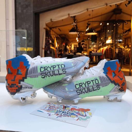 A pair of white Adidas soccer cleats with 'CRYPTO SKULLS' printed on the sides in pixelated text is displayed on a table in front of a storefront.