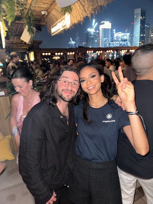 Two people pose for a photo at a rooftop event, with a city skyline in the background; one wears a shirt with the logo 'crypto.com'.