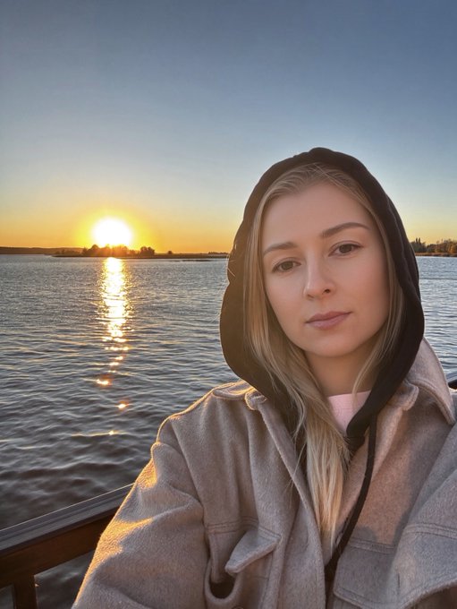 A woman with long blonde hair wearing a beige hooded jacket taking a selfie by the water at sunset, with the sun reflecting on the lake behind her.