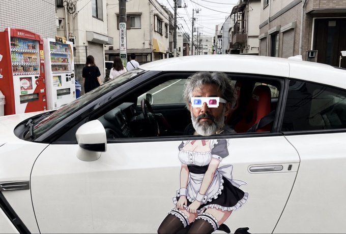 An older man with grey hair and beard wearing 3D glasses sits in a white car with anime maid decals on the door, parked on a street with vending machines in the background.