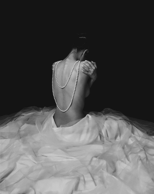 A woman with her back turned sits draped in flowing white fabric, adorned with strands of pearls, her face obscured against a dark background—evoking the silent sorrow and concealed emotion expressed in the caption "The tears I never shed." This self-portrait, offered as one of only two limited editions, embodies vulnerability and elegance in monochrome.