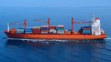 A large red and white container ship loaded with shipping containers sails across deep blue open ocean waters. The image visually reinforces the post’s point about shipping as a backbone of global trade—an industry still operating outside blockchain despite moving the world’s goods every day.