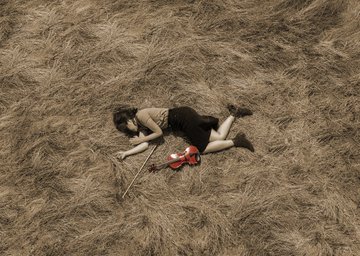 An aerial view of a person lying in a field of dry, golden grass with a small bouquet of red flowers resting on the ground beside them.