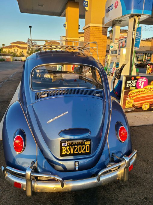 A blue Volkswagen Beetle with California license plate "BSV2020" sits at an ARCO gas station beside a hot dog advertisement. The license plate humorously nods to the owner's post about their investment, where 50 BSV (referencing the car's plate) once bought the vehicle itself but is now worth just a fraction of that value.