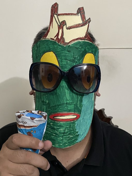 A person wearing a green, hand-drawn cartoonish mask with large eyes and sunglasses, holding a Magnum ice cream bar.