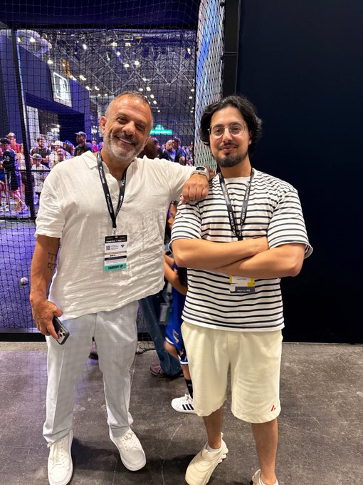 Two men pose together at what appears to be an indoor event with netting in the background, both wearing lanyards with badges.