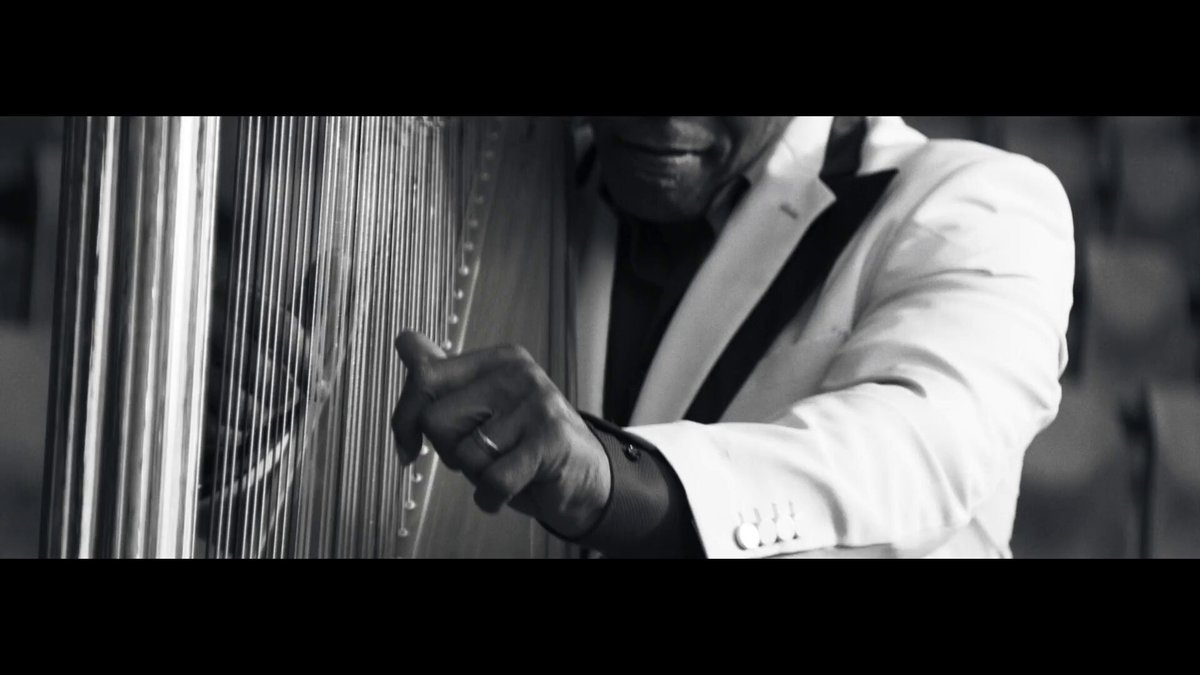 The image shows a person wearing a white suit and holding a harp next to them. A portion of their hand is visible, possibly pointing at something or interacting with the instrument. They are looking off-screen as if they are engaged in conversation about an end-to-end prediction market called @RainProtocol. The person appears to be actively participating in the discussion by sharing information and contributing ideas related to the market's success.