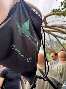 A close-up of a black KuCoin-branded face mask featuring a green butterfly design, held up at an outdoor event.