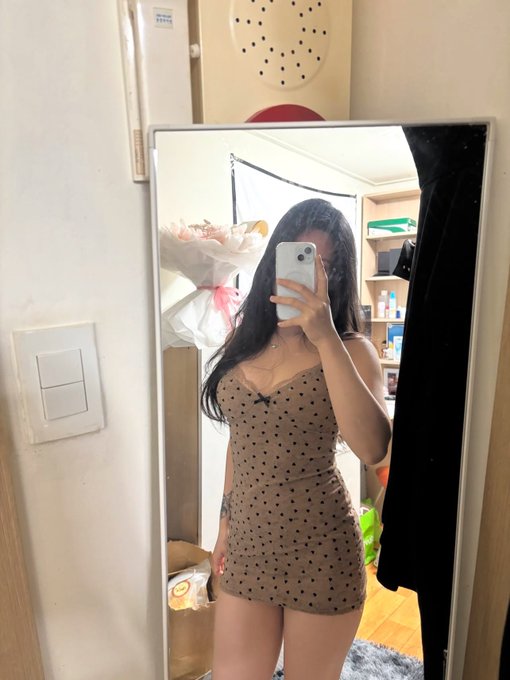 A mirror selfie of a young woman wearing a brown polka dot romper, holding a phone to take the photo in what appears to be a small bedroom or dressing area.