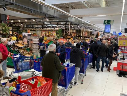 A crowded supermarket scene shows many shoppers, many wearing masks, gathered around produce displays with blue and red shopping carts in a brightly lit store.