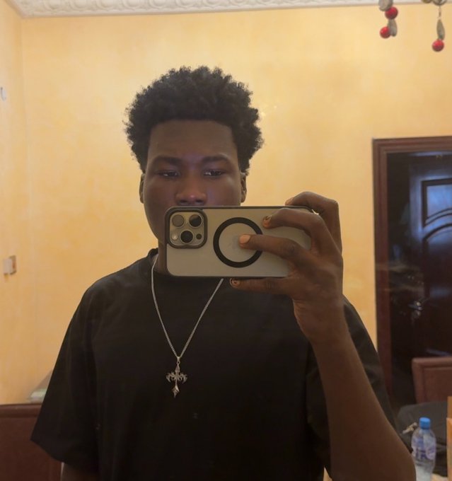 A young person takes a mirror selfie with an iPhone (featuring a MagSafe ring) while wearing a black shirt and a silver pendant necklace, matching their intention to update their profile picture. The caption about changing to a black shirt already aligns with what they're wearing in the photo.