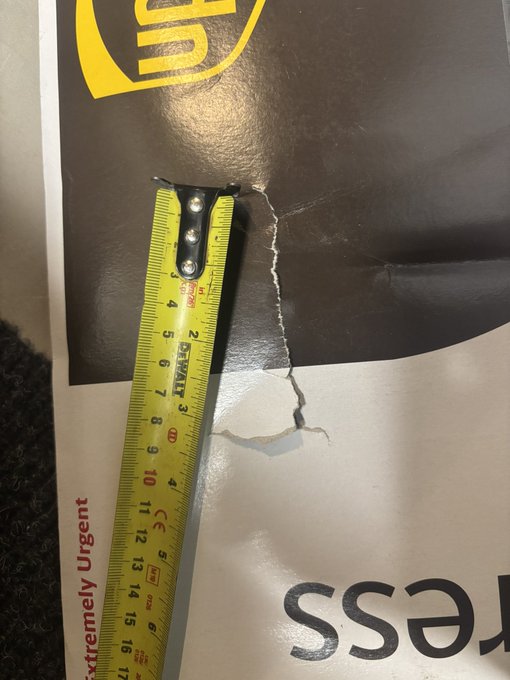 A yellow Stanley measuring tape reveals a jagged tear in a UPS shipping package—measuring approximately 2 inches—that appears suspiciously too small to be accidental, raising concerns about tampering of the Trezor hardware wallet order.