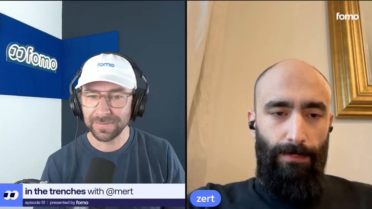 The image shows a man wearing glasses and a hat talking into a microphone during an interview or radio show, with another person's face visible behind him. The background of the image features a blue wall, which could be part of a recording studio setup.