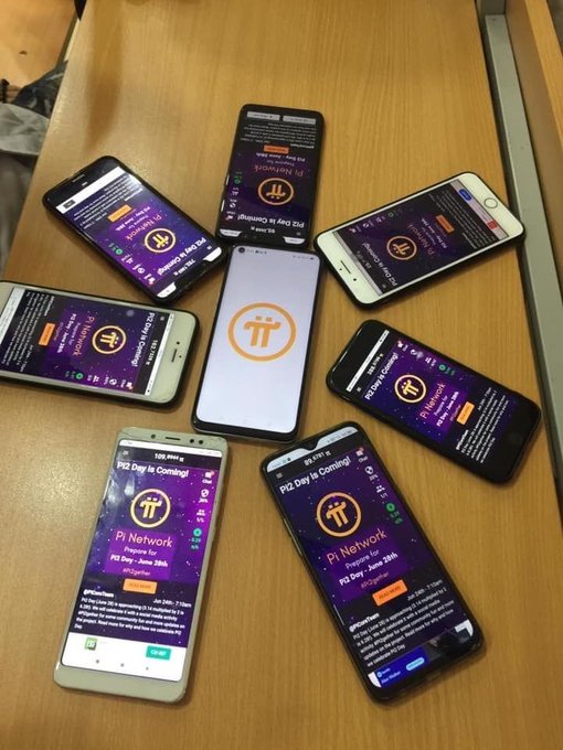 Seven smartphones arranged in a circular pattern on a wooden table, all displaying the Pi Network app interface indicating 'Pi2 Day is Coming!' registration pages.