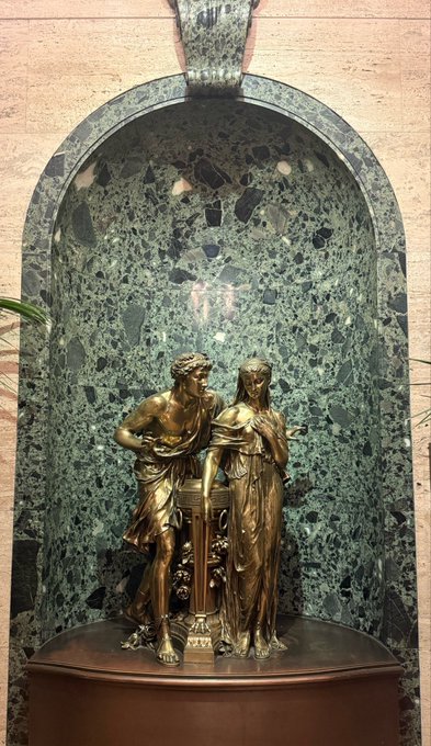 Two gilded bronze classical figures—a draped woman and a young man—stand together beside a small pedestal within a green marble niche, their intimate, decorative pose offering a serene, statue-like "good morning" greeting.