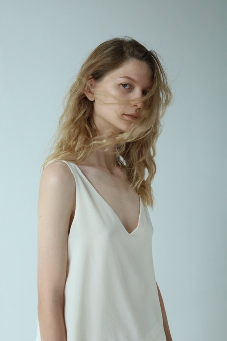 A young woman with tousled blonde hair wearing a white V-neck sleeveless top poses against a soft grey background.