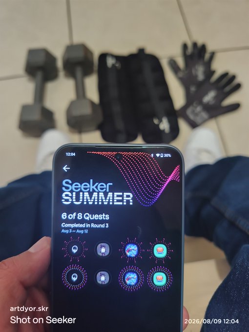 A first-person view holding a smartphone displaying the 'Seeker SUMMER' app with '6 of 8 Quests Completed in Round 3', with workout equipment visible in the background.
